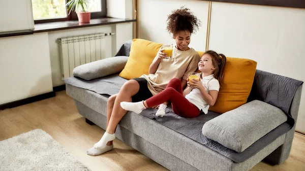 Aproveita o momento. afro-americana baby sitter divertido caucasiano bonito menina. Eles estão sentados no sofá, bebendo suco enquanto assistem TV — Fotografia de Stock