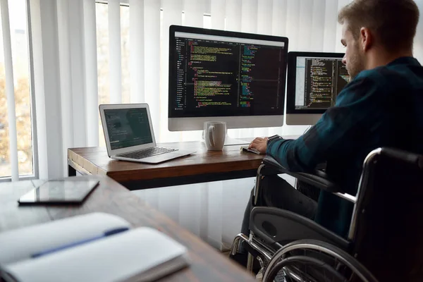 Ik kan met elke code werken. Geconcentreerde mannelijke web developer in een rolstoel schrijven programmacode op meerdere computerschermen tijdens het zitten op zijn werkplek in het moderne kantoor — Stockfoto