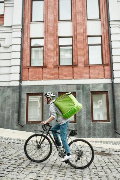 Junge Zustellerin mit Helm, Thermosack oder Rucksack, die mit dem Fahrrad durch die Stadt fährt und Essen ausliefert. Kurier, Lieferservice-Konzept — Stockfoto