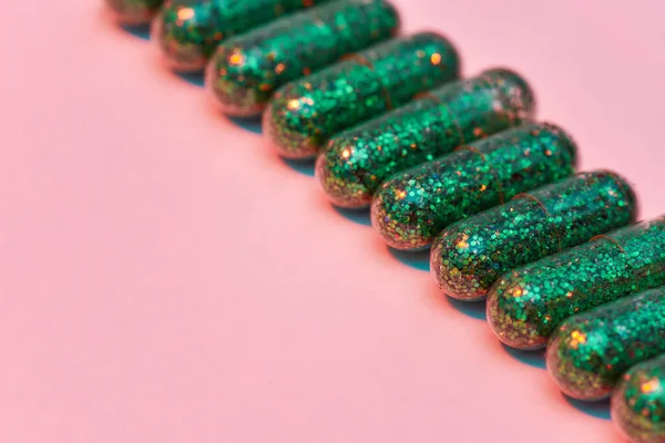 Tvůrčí koncept s mnoha zelenými třpytkami pilulky ležící diagonálně v řadě izolované na pastelově růžovém pozadí. Minimální styl, umělecký koncept — Stock fotografie