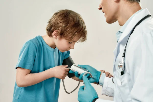 A trabalhar com crianças. Pediatra masculino profissional em uniforme médico medindo a pressão arterial de menino bonito contra fundo cinza. Saúde e medicina — Fotografia de Stock