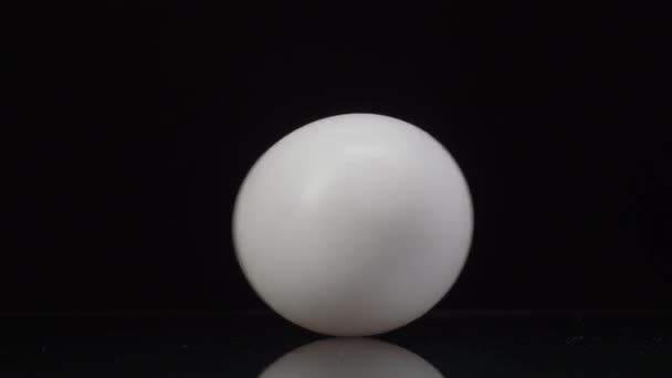 Huevo de gallina blanca girando y rodando sobre un fondo negro con hermoso reflejo. De cerca. — Vídeos de Stock