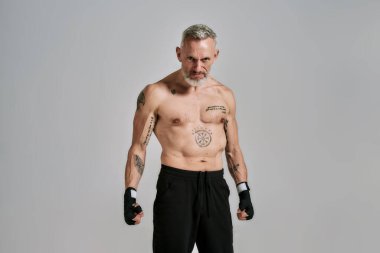 Yarı çıplak orta yaşlı atletik adam, kickboksçu sinirli görünüyor, agresif, gri arka planda stüdyoda duruyor. Muay Thai, Boks veya Kickboks konsepti