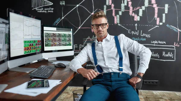 Geld laten groeien. Middelbare leeftijd Kaukasische handelaar in glazen kijken naar de camera terwijl zitten aan het bureau voor computer monitor. Schoolbord vol met grafieken en data-analyses op de achtergrond. — Stockfoto