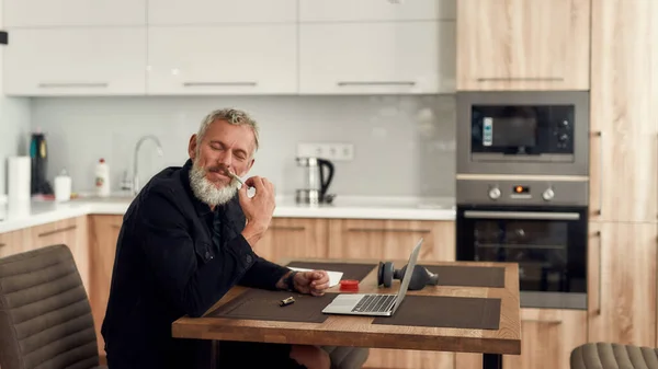 No Drama. Middle-aged man, artist enjoying the smell of marijuana cigarette or joint, sitting at home, in the kitchen and using laptop. Marijuana grinder, lighter and headphones on the table — Stock Photo, Image