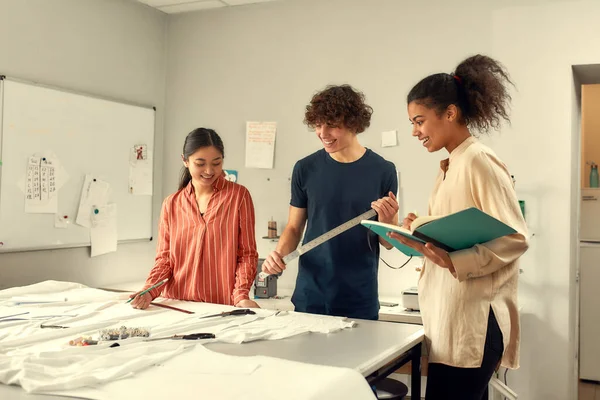 Be trendy for every time. Young male designer talking to female colleague while going to measure white fabric textile in a studio. Group of creative millennials working together — Stock Photo, Image