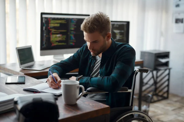 Zijaanzicht van jonge geconcentreerde mannelijke webontwikkelaar in een rolstoel die aantekeningen maakt terwijl hij op zijn werkplek zit met meerdere computerschermen op de achtergrond — Stockfoto