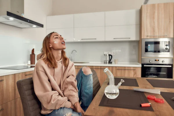 Relax your mind. Young caucasian woman exhaling the smoke while smoking marijuana from a bong or glass water pipe, sitting in the kitchen. Red marijuana grinder and lighter on the table