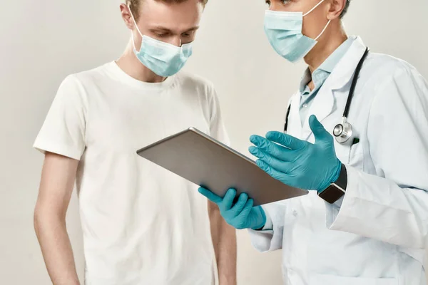 Doktor ve maskeli genç bir hasta. Tıbbi üniformalı yetişkin bir doktor elinde dijital tabletle genç bir adama teşhisi açıklıyor. — Stok fotoğraf