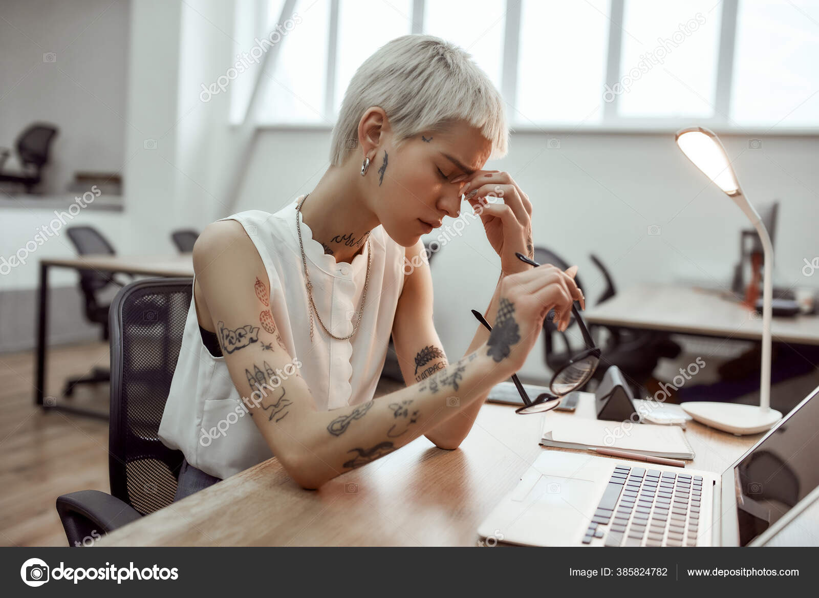 Feeling exhausted. Side view of frustrated young tattooed business woman  keeping eyes closed, holding her glasses in hand and massaging nose while  sitting at her working place in the modern office Stock