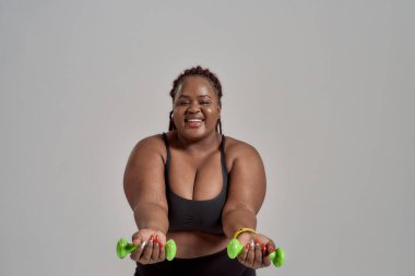 Doğru yönde. Plump, artı spor giyimli Afrikalı Amerikalı kadın kollarını öne uzatıyor, elinde yeşil dambıllar, gri arka planda stüdyoda poz veriyor.