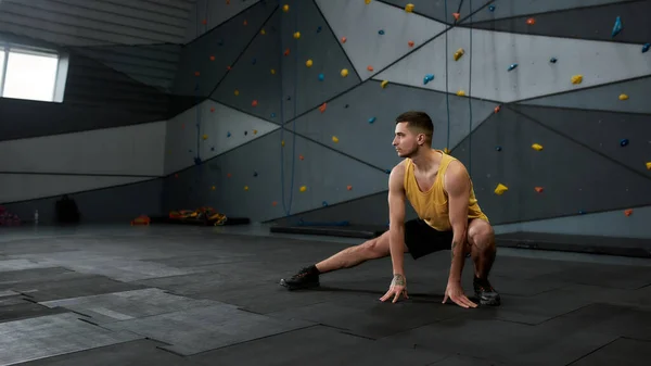 Tirador de cuerpo entero de joven activo en ropa deportiva mirando a un lado, mientras que estira el cuerpo contra la pared de escalada. Concepto de vida deportiva y escalada en roca —  Fotos de Stock