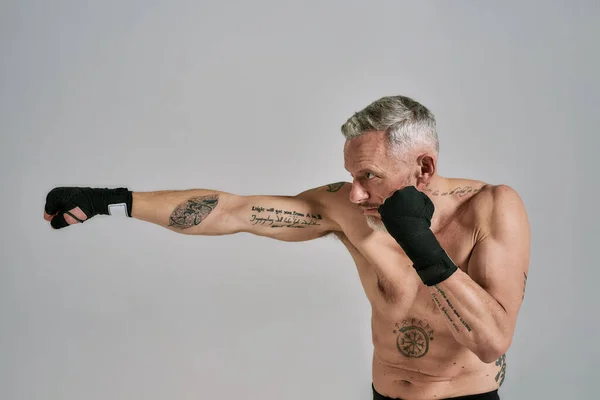 Apresúrate. Hombre atlético medio desnudo de mediana edad, kickboxer mirando a un lado mientras boxea, practicando golpes en el estudio sobre fondo gris. concepto de Muay Thai, Boxeo o Kickboxing —  Fotos de Stock