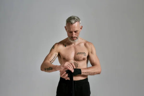 Mezza nuda uomo atletico di mezza età, kickboxer che avvolge le mani per Muay Thai, Boxe o Kickboxing, in piedi in studio su sfondo grigio — Foto Stock