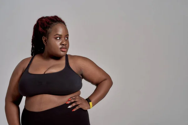 Fühlen Sie sich frei. Plump, plus size afrikanisch amerikanische Frau in schwarzer Sportbekleidung schaut beiseite, während sie im Studio vor grauem Hintergrund steht. Konzept des Sports, gesunder Lebensstil, positiver Körper, Gleichberechtigung — Stockfoto