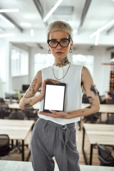 Look here. Young and attractive tattooed business woman with short haircut holding digital tablet and looking at camera while standing in modern coworking space
