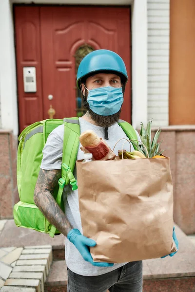 Hombre de reparto barbudo en casco con máscara y guantes debido a la aparición del virus Covid19, sosteniendo la bolsa de comestibles, mientras que la entrega de alimentos —  Fotos de Stock