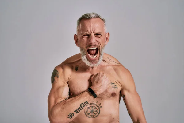 Inner power. Close up of half naked middle aged muscular man shouting at camera, beating his chest while posing in studio over grey background — Stock Photo, Image