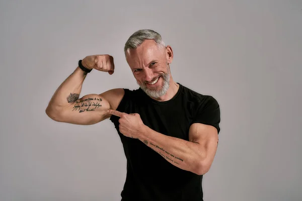 Titta på det här. Leende medelålders muskulös man i svart t skjorta visar sina biceps, tatueringar samtidigt poserar i studio över grå bakgrund — Stockfoto