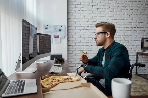 Çok lezzetli. Tekerlekli sandalyede pizza yiyen ve iş yerinde otururken birden fazla bilgisayar ekranına bakan beyaz erkek web geliştiricisinin yan görüntüsü. — Stok fotoğraf