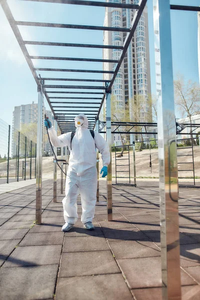 Neat cleaning. Sanitization and disinfection of the city due to the emergence of the Covid19 virus. Man in protective suit and mask at work near sports ground — Stock Photo, Image