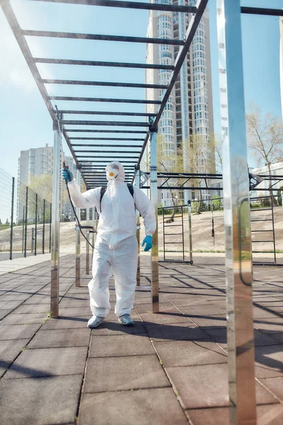 Snygg städning. Sanering och desinfektion av staden på grund av uppkomsten av Covid19-viruset. Man i skyddsdräkt och mask på jobbet nära idrottsplats — Stockfoto