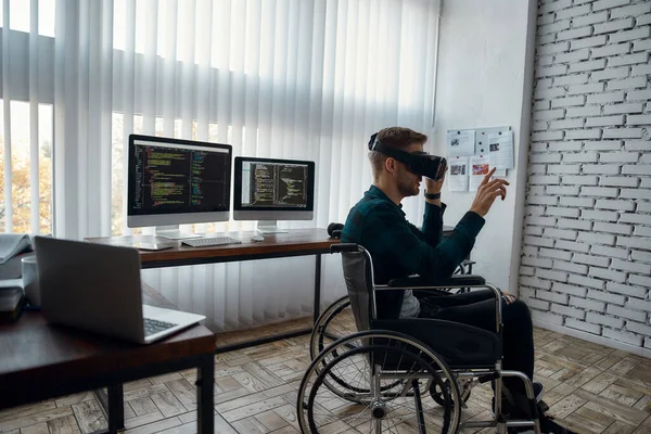 Zijaanzicht van een jonge mannelijke webontwikkelaar in een rolstoel met een virtual reality bril tijdens het werken op zijn werkplek in het moderne heldere kantoor. Meerdere computerschermen met programmacode — Stockfoto