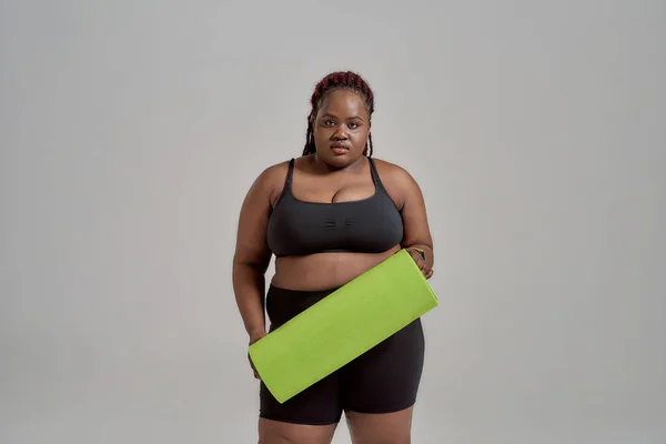 Plump, plus size afrikanisch amerikanische Frau in Sportbekleidung hält grüne Yogamatte für Fitness, posiert im Studio vor grauem Hintergrund — Stockfoto