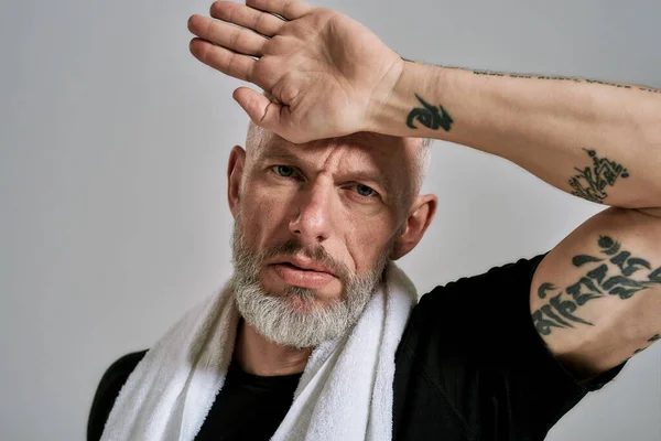 Uomo muscoloso di mezza età in t shirt nera guardando la fotocamera mentre asciuga il sudore sulla fronte dopo l'allenamento, in posa in studio su sfondo grigio — Foto Stock