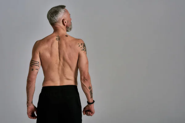 Meio nu homem musculoso de meia idade olhando para o lado, mostrando as costas, corpo enquanto posando em estúdio sobre fundo cinza — Fotografia de Stock