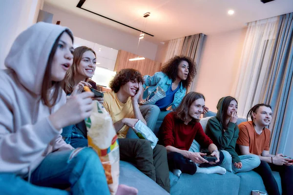 Οι φίλοι διασκεδάζουν στο σπίτι. Νέοι ευτυχισμένοι πολυπολιτισμικοί άνθρωποι που παίζουν βιντεοπαιχνίδια, τρώνε σνακ και χαλαρώνουν μαζί στον καναπέ στο σπίτι — Φωτογραφία Αρχείου