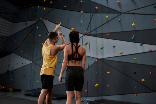 Altitud, no actitud. Mujer joven activa en ropa deportiva va a subir, instructor masculino ayudándola, de pie contra la pared de escalada de entrenamiento artificial. Concepto de vida deportiva y escalada en roca —  Fotos de Stock