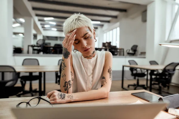 Terrible headache. Young tattooed business woman keeping eyes closed, touching her head, suffering from the headache while sitting at her working place in the modern office