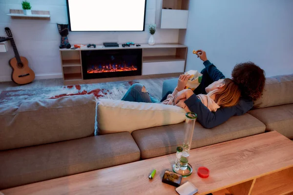 Vue arrière du jeune couple regardant la télévision ensemble, mangeant des collations après avoir fumé de la marijuana d'un bong ou d'un tuyau d'eau en verre. Ils se détendent sur le canapé le soir à la maison — Photo