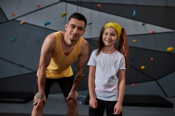Genç erkek eğitmen ve aktif küçük kız kameraya bakıyor, yapay tırmanma duvarına karşı duruyor. Spor hayatı ve kaya tırmanışı kavramı — Stok fotoğraf
