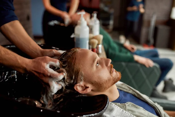 Вид сбоку на молодого красивого рыжего мужчину, опирающегося на раковину, пока парикмахер моет волосы перед стрижкой — стоковое фото
