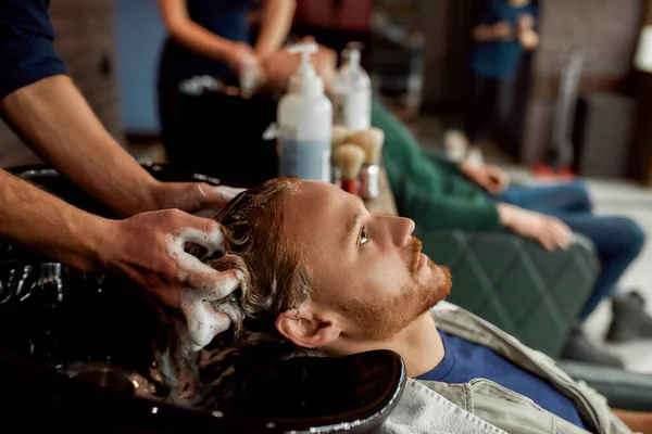 Zijaanzicht van een jonge knappe roodharige man leunend op de gootsteen terwijl de kapper zijn haar waste voor een kapsel — Stockfoto