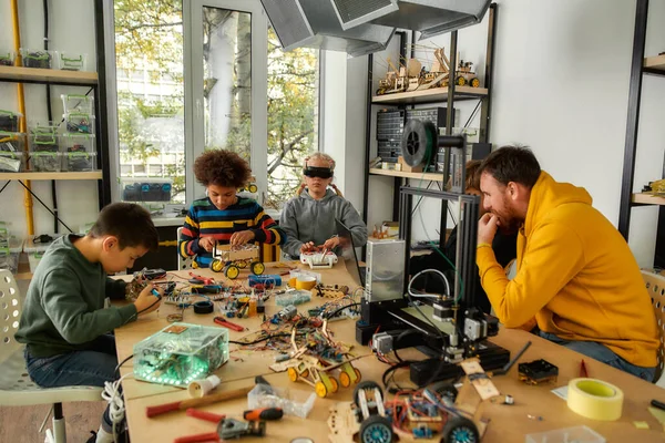 Learning, friendship and fun. Young technicians building robots and vehicles, using soldering iron to join chips and wires, testing toys together with a male teacher at a stem robotics class — Stock Photo, Image