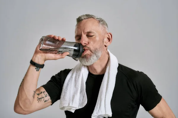 Återfukta. Medelålders muskulös man i svart t skjorta stänga ögonen medan dricksvatten från flaskan, poserar i studio över grå bakgrund — Stockfoto