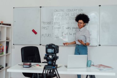 Çevrimiçi ders veriyorum. Genç afro amerikan bayan İngilizce öğretmeni beyaz tahtanın yanında duruyor ve gülümsüyor, İngilizce dilbilgisi kurallarını online olarak açıklıyor.