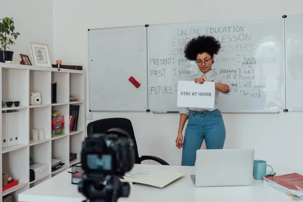 Online tanterem. Fiatal afro-amerikai angol tanárnő bemutató papírt szöveggel HOME otthon webkamera, miközben ellen fehér tábla — Stock Fotó