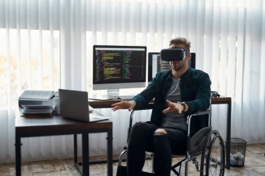 VR iş başında. Modern ofisindeki iş yerinde çalışırken tekerlekli sandalyede sanal gerçeklik gözlüğü takan genç bir erkek ağ geliştirici. Yüksek Teknoloji