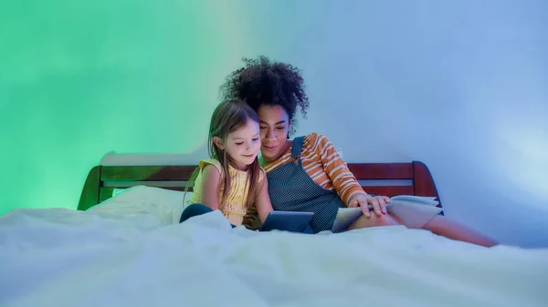 The proper childcare. African american woman baby sitter and cute little girl reading book fairy tale and using tablet pc in bed. Kid and nanny in bedroom in the evening — Stock Photo, Image