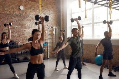 Daha güçlü. Daha hızlı! Endüstriyel spor salonunda spor yaparken dambılı kaldıran bir grup insan. Grup eğitimi, takım çalışması konsepti