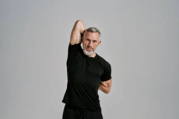 Uomo muscoloso di mezza età in abbigliamento sportivo nero guardando la fotocamera mentre riscalda il suo corpo prima dell'allenamento in studio su sfondo grigio — Foto Stock