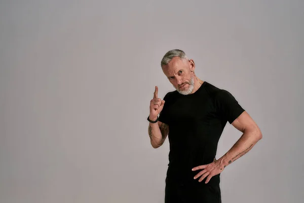 Uomo di mezza età in t shirt nera guardando la fotocamera, che punta verso l'alto mentre posa in studio su sfondo grigio — Foto Stock