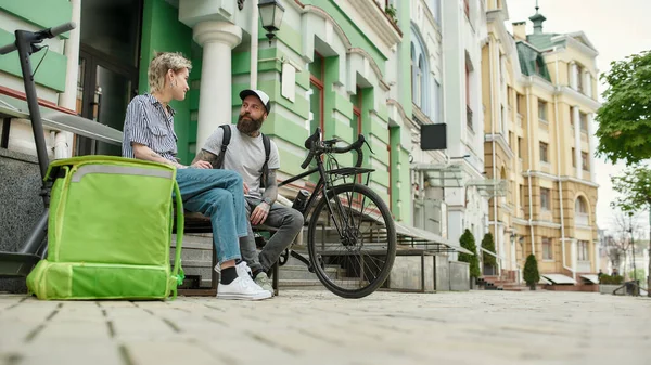 二つの陽気な宅配便、若い男と女性がベンチに座って、食べ物や製品を配達しながら屋外で話して、スクーターや自転車を使用して — ストック写真