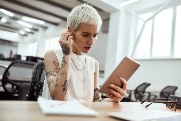 Rozmowa przez Skype. Portret młodej uroczej blondynki wytatuowanej bizneswoman zakładającej bezprzewodowe słuchawki i korzystającej z touchpada podczas siedzenia w nowoczesnym biurze — Zdjęcie stockowe