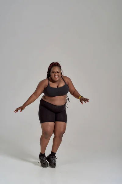 Ich liebe mich selbst. In voller Länge Aufnahme von molligen, plus size afrikanisch-amerikanische Frau in Sportbekleidung Spaß haben, tanzen im Studio vor grauem Hintergrund — Stockfoto