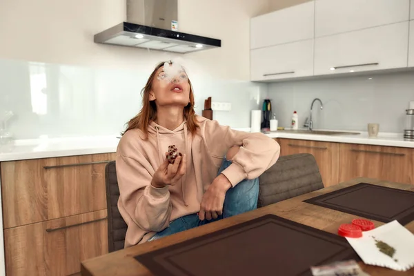 Feeling Good. Young caucasian woman exhaling the smoke while smoking marijuana from a glass pipe, sitting in the kitchen. Red weed grinder on the table